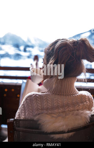 Junge hübsche Dame in gemütlichen Pyjama sitzen draußen auf der Terrasse und trinken Kaffee. Panoramablick vom Hotel auf der Winter Schweiz Doppelzi. Stockfoto