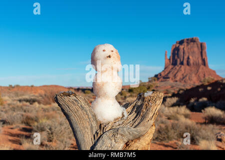 Close Up. Kleiner Schneemann auf toten Baumstamm unter blauem Himmel. Verschwommen wüste Landschaft mit roten Sandstein butte im Hintergrund. Stockfoto