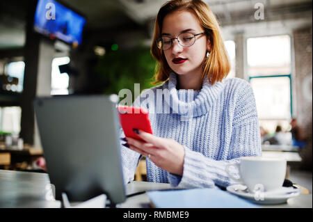 Fröhliche junge schöne rothaarige Frau in Gläser mit Ihrem Telefon, Touchpad, Notebook, wenn an Ihrem Arbeitsplatz sitzen auf Cafe mit Tasse Kaff Stockfoto