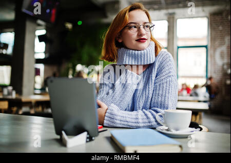 Fröhliche junge schöne rothaarige Frau in Gläser mit Ihrem Telefon, Touchpad, Notebook, wenn an Ihrem Arbeitsplatz sitzen auf Cafe mit Tasse Kaff Stockfoto