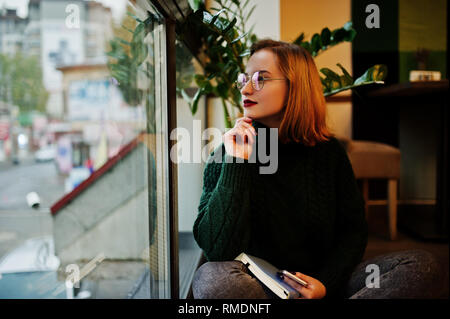 Träumt etwas schönes. Fröhliche junge schöne rothaarige Frau in Gläser, grüne warmen Wollpullover, mit Ihrem Notebook, während der Sitzung Stockfoto