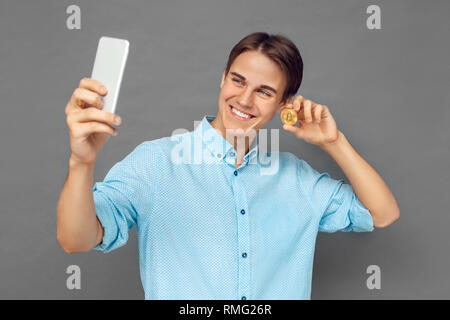 Junge Mann isoliert auf graue Wand unter selfie Foto auf dem Smartphone mit cryptocurrency Münze lächelt glücklich Stockfoto