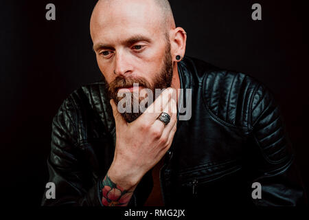Nachdenklich kahlen Hipster in Leder Jacke mit Tattoos auf Hand kratzen Bart auf schwarzem Hintergrund Stockfoto