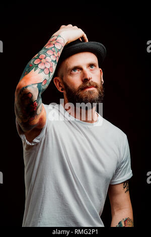 Fröhliche bärtigen Hipster in Hut und t-shirt mit Tattoos auf Arme auf schwarzem Hintergrund Stockfoto
