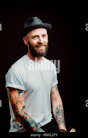 Fröhliche bärtigen Hipster in Hut und t-shirt mit Tattoos auf Händen auf schwarzem Hintergrund Stockfoto