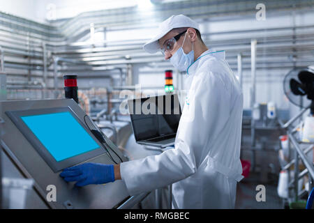 Mann in einem weißen Gewand und einer Maske mit einem Laptop in der Hand steht in der Nähe der digitalen Bildschirm im Werk Stockfoto