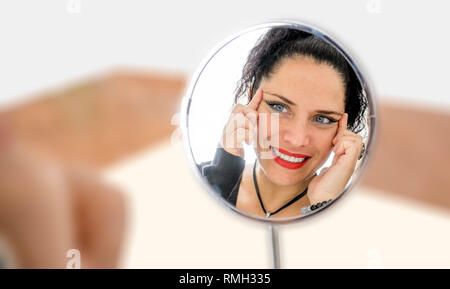 Im mittleren Alter normale Frau sich selbst in einem Spiegel Anti - Aging creme & lächelnd mit roten Lippen und blauen Augen. Sommersprossen Gesicht haut. Isoliert Stockfoto