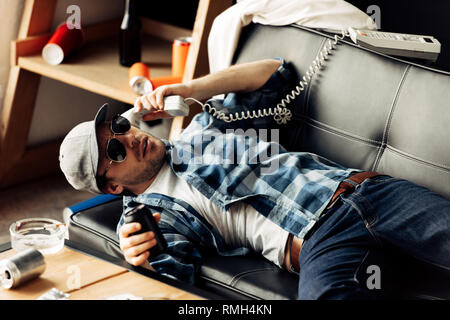 Schöner Mann, Sonnenbrille und Kappe liegen auf dem Sofa und Sprechen auf retro Telefon im Wohnzimmer Stockfoto