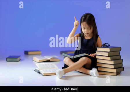 Süße kleine Mädchen im Kleid ist lesen Buch, nach oben zeigt und die Idee während der Sitzung zwischen Büchern. Kind und eine Menge Bücher. Kinder und Bildung. Stockfoto