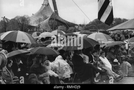 Besucher mit Sonnenschirmen im Regen im Olympiastadion sitzen und die Münchner Evangelium Sound Chor Konzert ansehen. Stockfoto