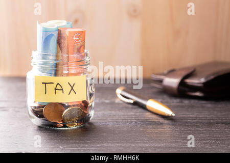 Glas Glas mit Münzen und Euro mit der Aufschrift Steuer auf einem schwarzen Holztisch und Hintergrund. Stift und Lederetui. Konzept der finanziellen Einsparungen t Stockfoto