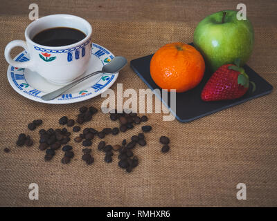 Frühstück eines Porzellan Tasse Kaffee, Kaffeebohnen, einen alten Löffel und eine Platte mit ein grüner Apfel, eine Mandarine und eine Erdbeere auf einem vintage Sackleinen Stockfoto