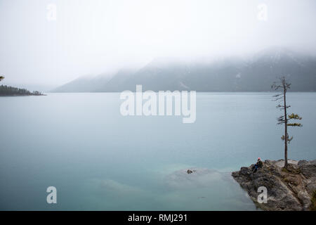 Ein Mädchen in einem rosa Mütze sitzt und blickt auf Lake Minnewanka an einem nebligen Tag in Banff National Park, Kanada Stockfoto