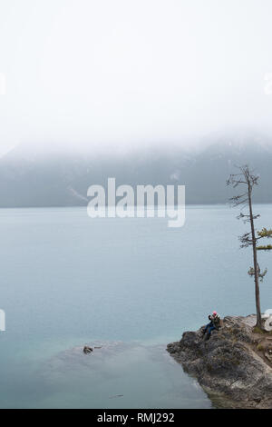 Ein Mädchen in einem rosa Mütze sitzt und blickt auf Lake Minnewanka an einem nebligen Tag in Banff National Park, Kanada Stockfoto