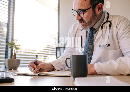 Arzt schreibt in seinem Tagebuch sitzend an seinem Schreibtisch. Männliche reife Arzt schreiben Hinweise für Patienten in der Klinik. Stockfoto