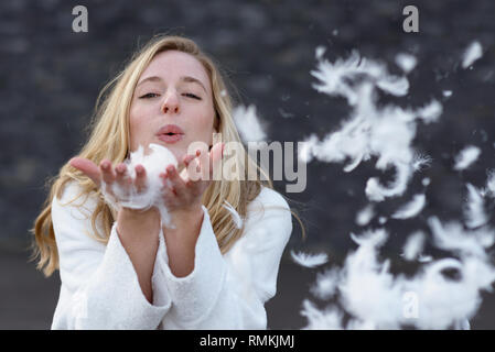 Spaß verspielte junge Frau bläst weiße Federn auf die Kamera zu einem Stapel in die Hände, als sie draußen auf einem grauen Hintergrund mit Kopie Spa steht Stockfoto