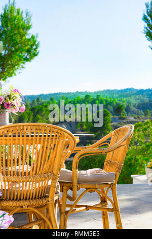 Terrasse von luxuriöse Villa mit Korbstühlen Tabelle Blumen Blumenstrauß auf dem Tisch faszinierende Aussicht auf Tal in den Bergen mit grünen Bäumen bedeckt. Sunn Stockfoto