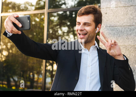 Foto von bärtigen Executive Manager im Anzug holding Handy für selfie während im Freien stehend gegen Gebäude Stockfoto