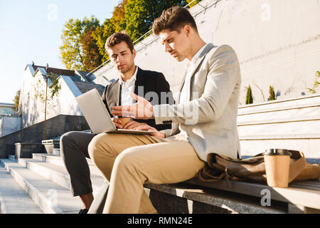 Foto von europäische Geschäftsleute in Anzügen sitzen auf der Treppe im Freien und mit Laptop während der sonnigen Tag Stockfoto