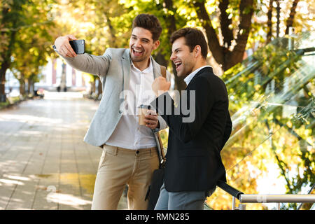 Foto des erfolgreichen Business Männer in Anzügen mit Smartphone beim Gehen im Freien durch die grünen Park mit Kaffee zum Mitnehmen während der sonnigen Tag Stockfoto