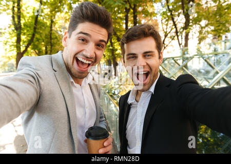 Foto der kaukasischen sachlich Jungs in Anzügen, die selfie Foto auf dem Smartphone während Outdoor wandern durch grüne Park mit Kaffee zum Mitnehmen Stockfoto