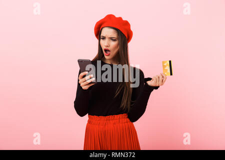 Porträt eines zornigen schöne junge Frau das Tragen der roten Baskenmütze stehend über rosa Hintergrund isoliert, an Handy suchen, Holding plastik Kredit c Stockfoto