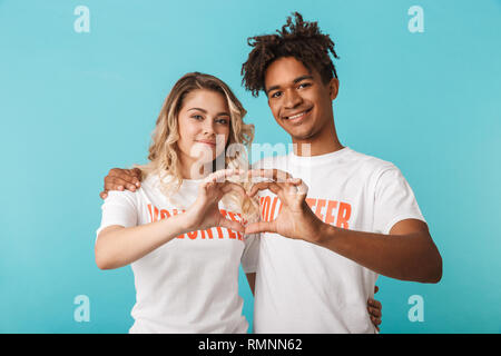 Gerne zuversichtlich multiethnischen Paar tragen Helfer t-shirt stehend über blauen Hintergrund isoliert, Herz Geste Stockfoto