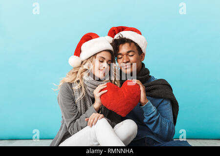 Happy multirassischen Junges Paar Tragen Weihnachten rote Hüte über blau Hintergrund isoliert, sitzend, mit roten Herzen Stockfoto