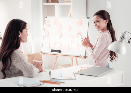 In der Nähe der Tafel. Lächelnd clever Schülerin zeigt auf einer Tafel Übergabe einen Stift in den Händen vor der fürsorglichen Mutter steht Stockfoto