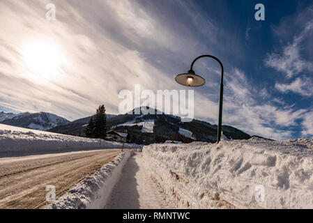 Straßenlaterne auf während des Tages auf eine verschneite, leere Straße im Winter. Sonne und Berge im Hintergrund. Rohrmoos Untertal, Schladming Dachstein Österreich Europa Stockfoto