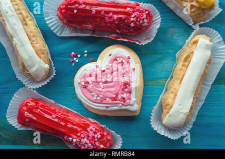 Eclairs mit vanila, Himbeer und Mango Aromen und Rosa herzförmige Plätzchen auf Blau Holz- Hintergrund Stockfoto