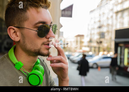 Junger stattlicher Mann mit grünen Kopfhörer und Sonnenbrille genießen eine Zigarette auf der Straße. Stockfoto