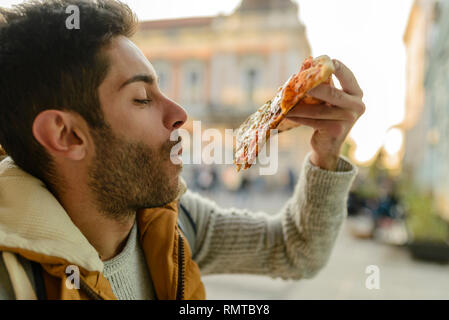 Gut aussehender bärtiger junger Mann mit orange Jacke essen Pizza in der Stadt. Stockfoto