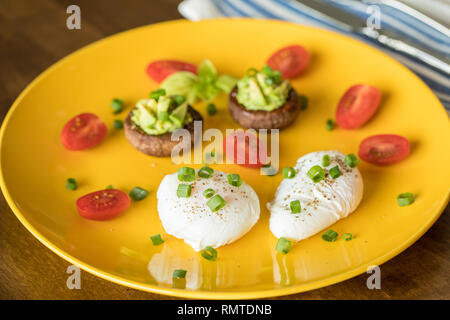 Pochierte Eier mit Avocado gefüllte Champignons, Tomaten und grüne Zwiebeln auf einer fröhlichen gelben Schild an einem Holztisch Stockfoto