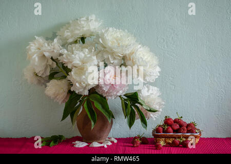 Einen großen Blumenstrauß von Pfingstrosen in Vase aus Ton und eine Schüssel Erdbeeren auf einem Baum. Romantische Stimmung. Stockfoto