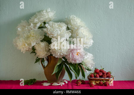 Einen großen Blumenstrauß von Pfingstrosen in Vase aus Ton und eine Schüssel Erdbeeren auf einem Baum. Romantische Stimmung. Stockfoto