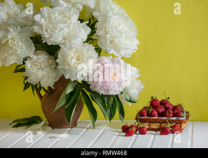 Einen großen Blumenstrauß von Pfingstrosen in Vase aus Ton und eine Schüssel Erdbeeren auf einem Baum. Romantische Stimmung. Stockfoto