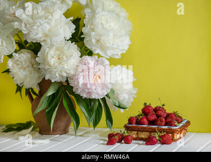 Einen großen Blumenstrauß von Pfingstrosen in Vase aus Ton und eine Schüssel Erdbeeren auf einem Baum. Romantische Stimmung. Stockfoto