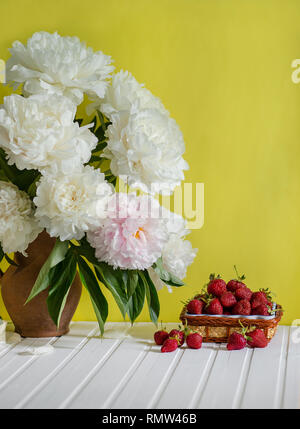 Einen großen Blumenstrauß von Pfingstrosen in Vase aus Ton und eine Schüssel Erdbeeren auf einem Baum. Romantische Stimmung. Stockfoto