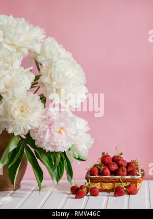 Einen großen Blumenstrauß von Pfingstrosen in Vase aus Ton und eine Schüssel Erdbeeren auf einem Baum. Romantische Stimmung. Stockfoto