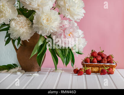 Einen großen Blumenstrauß von Pfingstrosen in Vase aus Ton und eine Schüssel Erdbeeren auf einem Baum. Romantische Stimmung. Stockfoto