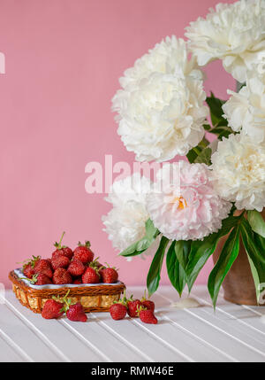 Einen großen Blumenstrauß von Pfingstrosen in Vase aus Ton und eine Schüssel Erdbeeren auf einem Baum. Romantische Stimmung. Stockfoto