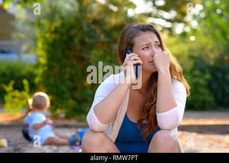 Betonte gedrückt junge Mutter reden, und auf einem Mobiltelefon, als sie draußen sitzt auf einem Spielplatz mit ihrem Baby Sohn hinter ihr Spielen Stockfoto