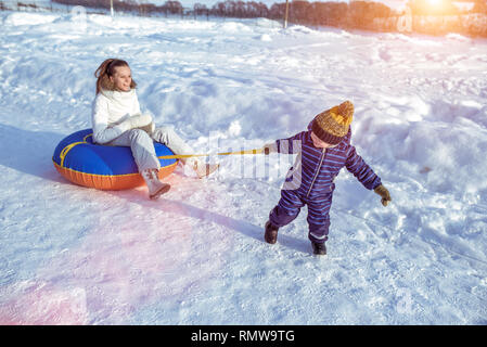 Ein kleiner Junge schleppt ein schlauchsystem Seil auf Seil, an dem die Mutter sitzt. Gerne spielen in der Natur im Winter. Stockfoto