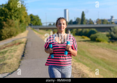 Passen junge Frau joggen kleine Hantel Gewichte in den Händen entlang einer ländlichen Straße glücklich lächelnd, als sie nähert sich die Kamera beim Genießen Ihrer Stockfoto