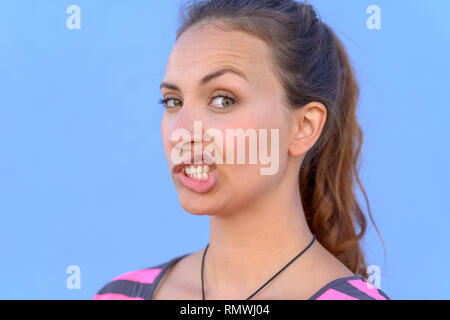 Attraktive junge Frau zieht eine lustige Ausdruck auf eine Seite ihr Gesicht über einen blauen Himmel Hintergrund in einer Nahaufnahme 7/8 Kopf geschossen Stockfoto