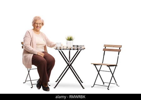 In voller Länge Porträt einer älteren Frau an einem Tisch sitzen mit einer Tasse Kaffee und lächelnd in die Kamera auf weißem Hintergrund Stockfoto