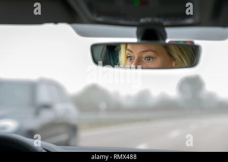 Frau, die ein Auto auf einer Landstraße an einem nebligen Tag mit ihren Augen weiß in den Spiegel geöffnet wider Stockfoto