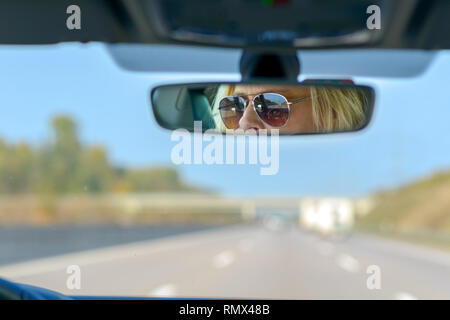 Frau, der ein Auto fährt auf einer Autobahn mit einem Blick nach vorne durch die Windschutzscheibe und ihre Gläser in den Rückspiegel reflektiert Stockfoto