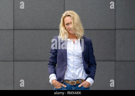Attraktive ernste trendy blonde Frau, mit den Händen in den Taschen und Haare vor ihrem Gesicht fallen Suchen nachdenklich an der Kamera Stockfoto